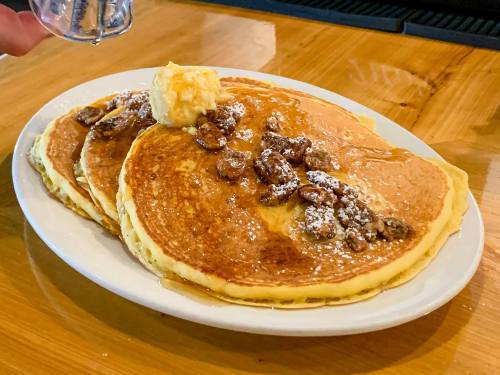 A plate of fluffy pancakes topped with butter, nuts, and a drizzle of syrup on a wooden table.
