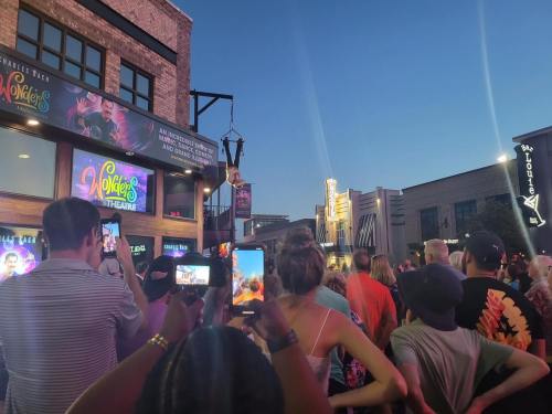 A crowd gathers outside a theater, watching a performance as people capture the moment on their phones at dusk.