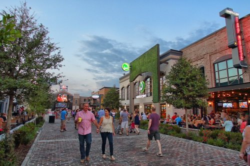 A couple walks hand-in-hand along a lively street lined with restaurants and outdoor seating at dusk.
