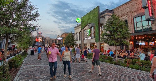 A lively outdoor scene with people walking, dining, and socializing in a vibrant shopping area at dusk.