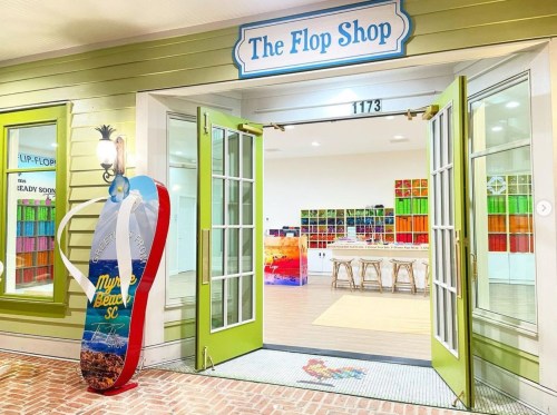 Brightly lit entrance of "The Flop Shop" with open doors, colorful decor, and a surfboard sign outside.