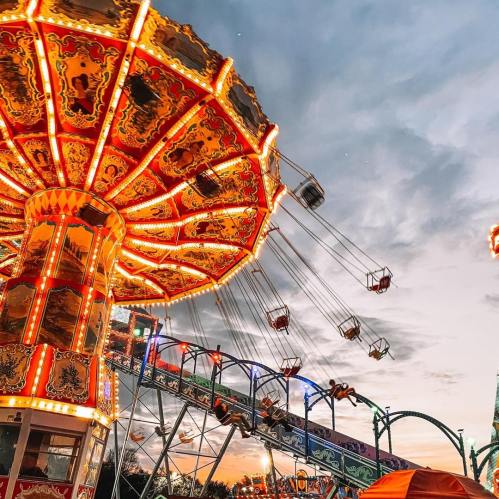 A vibrant carnival scene at sunset, featuring a colorful swing ride and a roller coaster illuminated by lights.