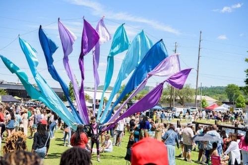 A vibrant outdoor festival scene with colorful fabric banners and a large crowd enjoying the festivities under a clear sky.
