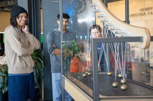 Three young men observe a Newton's cradle exhibit in a science center, smiling and engaged with the display.