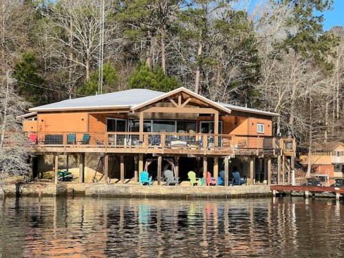 A wooden lakeside house with a large deck, surrounded by trees, reflecting in the calm water below.