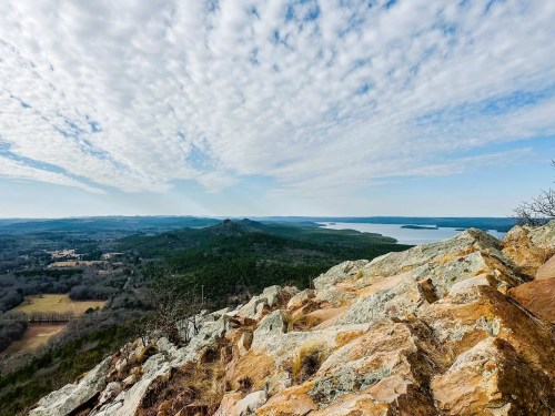 A panoramic view from a rocky summit, showcasing rolling hills, a lake, and a cloudy sky.