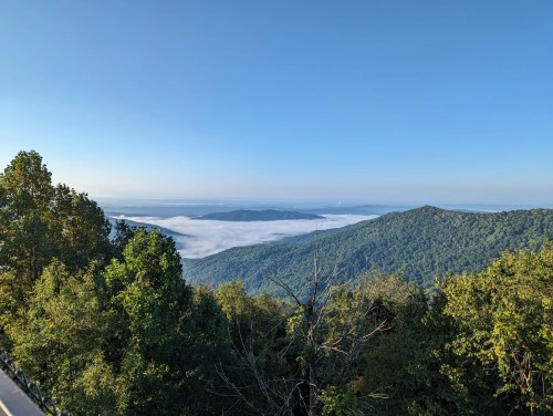 A scenic view of mountains covered in fog under a clear blue sky, with lush green trees in the foreground.