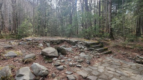 A stone pathway leads through a forested area with moss-covered rocks and steps, surrounded by trees and greenery.