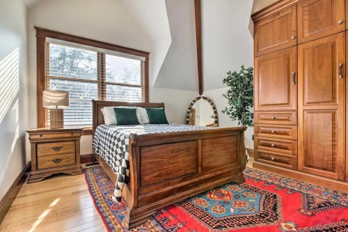 Cozy bedroom featuring a wooden bed, nightstand, large wardrobe, and a colorful rug under natural light from windows.