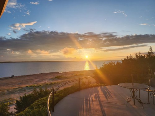 Sunset over a calm lake, with clouds and a golden sky, viewed from a patio with a table and chairs.