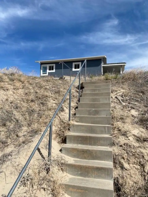 A set of concrete stairs leads up a sandy hill to a blue house under a clear blue sky.