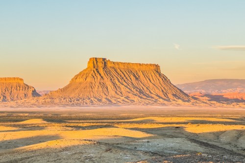A golden-hued mesa rises against a clear sky, surrounded by a vast, arid landscape at sunset.