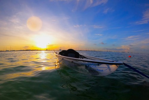A kayak floats on calm water at sunset, with a colorful sky and distant boats in the background.