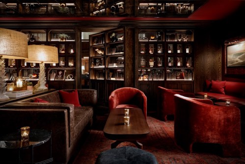 Cozy, dimly lit lounge with plush red seating, wooden shelves filled with books, and warm lighting.
