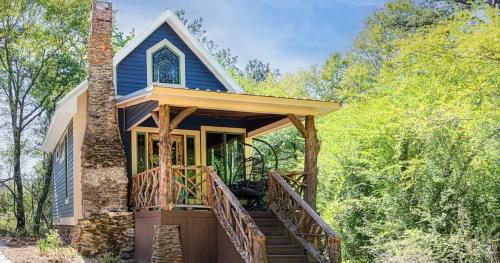 A charming blue cottage with a stone chimney, surrounded by greenery and featuring a cozy porch.