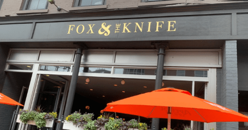 Exterior of a restaurant named "Fox & The Knife," featuring orange umbrellas and potted plants.