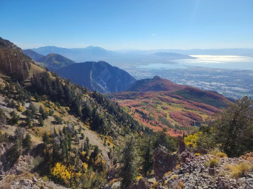 A panoramic view of colorful autumn foliage on mountains overlooking a valley and lake under a clear blue sky.