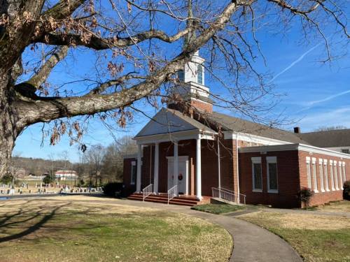 A brick church with a steeple, surrounded by a grassy area and bare trees, under a clear blue sky.