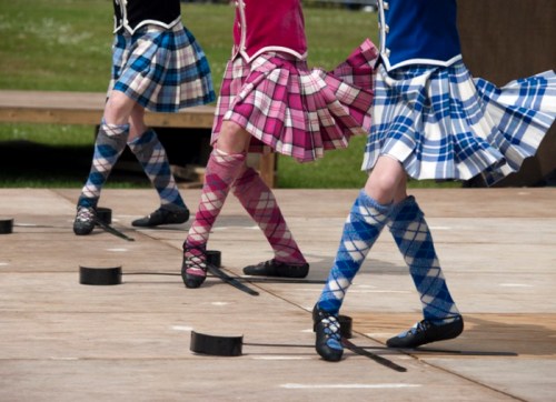 Three dancers in colorful kilts and knee-high socks perform on a wooden stage, showcasing traditional Scottish dance.