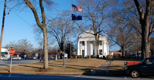 A small town scene featuring a white building, American and state flags, trees, and a stop sign on a clear day.
