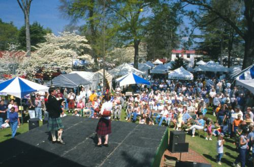 A lively outdoor festival scene with performers in kilts on stage and a large crowd enjoying the event under blue skies.
