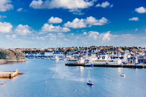 A scenic view of a harbor with boats, surrounded by charming buildings and a blue sky with fluffy clouds.