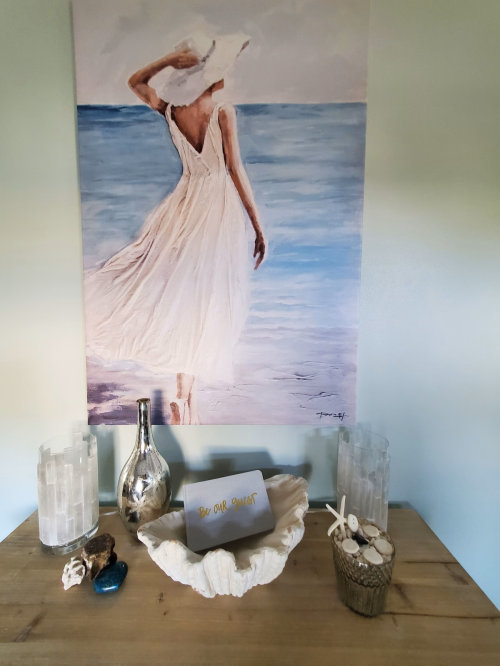 A serene beach-themed decor featuring a painting of a woman in a white dress, with decorative items on a wooden table.