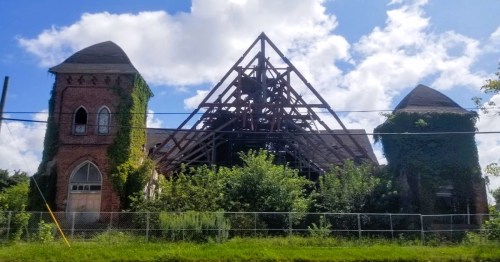 Abandoned brick building with two towers, overgrown with vines, under a blue sky with scattered clouds.