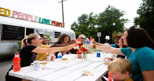 A diverse group of people joyfully toasting with drinks at a picnic table, surrounded by food and a food truck.