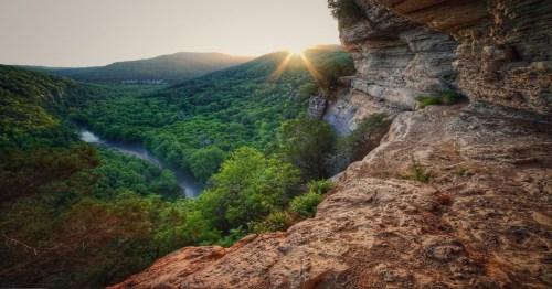 Sunset over a lush green valley, with a river winding through and rocky cliffs in the foreground.
