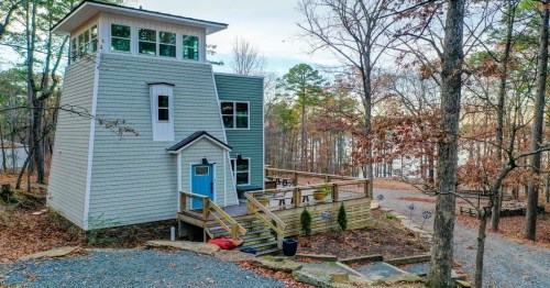 A modern, multi-story house with a blue door, surrounded by trees and a gravel path near a lake.