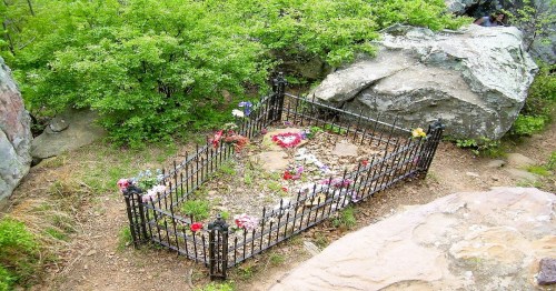 A small, fenced grave adorned with flowers, surrounded by rocks and greenery in a natural setting.