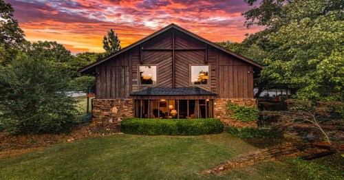 A rustic wooden house with stone accents, surrounded by greenery, under a vibrant sunset sky.