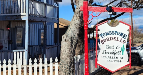 Sign for "Tombstone Bordello Bed & Breakfast" hanging from a tree, with a white picket fence and a historic building in the background.