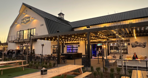 A modern barn-style building with outdoor seating, string lights, and a lively atmosphere during sunset.