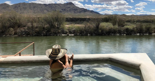 A person relaxes in a pool by a river, with mountains and clouds in the background.