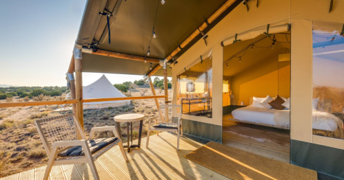 A cozy glamping tent with a wooden deck, featuring a bed, seating area, and scenic views of nature.