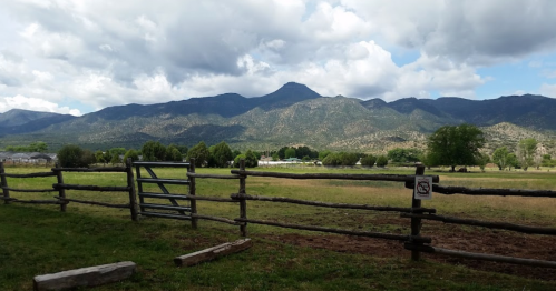 A scenic view of mountains under a cloudy sky, with a grassy field and wooden fence in the foreground.