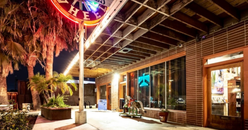 A vibrant storefront with neon lights, palm trees, and a playful horse ride outside, set in a lively evening atmosphere.