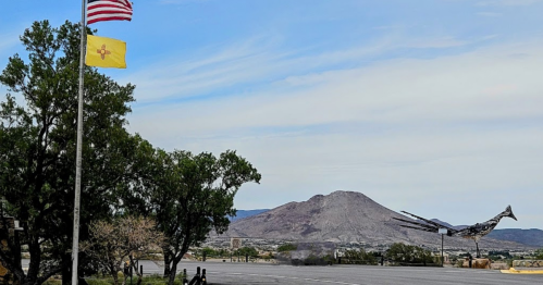 A U.S. flag and New Mexico flag fly near a mountain, with a sculpture of a bird in the foreground.