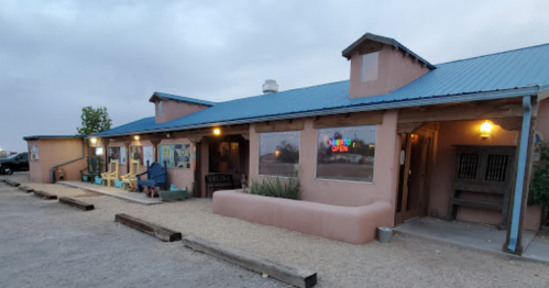 A rustic building with a blue roof, open sign, and outdoor seating, set against a cloudy sky.