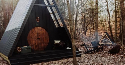 A black A-frame cabin in a wooded area, with a round wooden door and a fire pit nearby. Autumn leaves cover the ground.