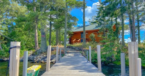 A wooden dock leads to a log cabin surrounded by tall trees and a clear blue sky.