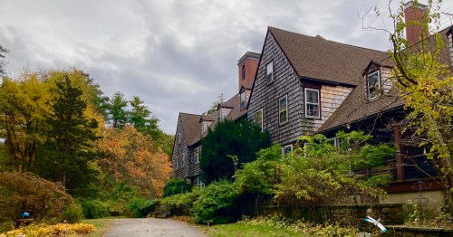 A large, rustic house surrounded by trees and autumn foliage under a cloudy sky.