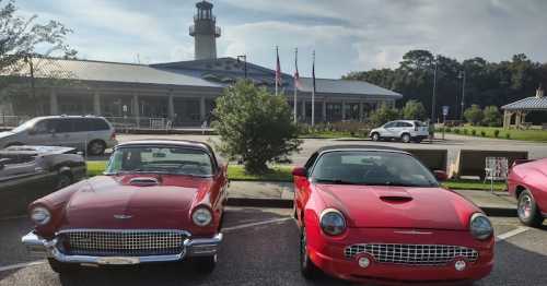 Two red classic cars parked in front of a building with a lighthouse, surrounded by trees and flags.