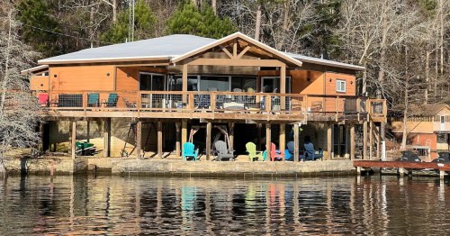 A lakeside cabin with a large deck, colorful chairs, and a serene water view surrounded by trees.