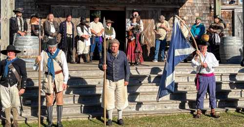 A group of people in historical costumes stands on a stage, some holding flags and instruments, in a festive outdoor setting.