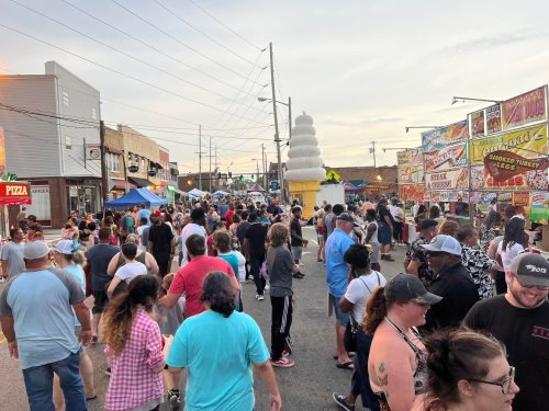 A crowded street fair with people walking among food stalls and colorful banners under a clear sky.