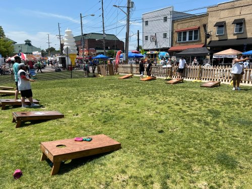 A sunny outdoor scene with people playing cornhole on a grassy area, surrounded by tents and shops.