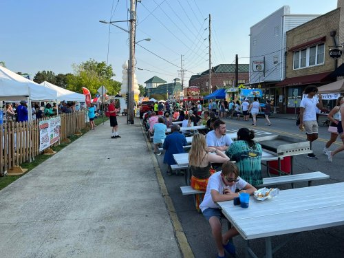 A lively street festival with people dining at picnic tables, food vendors, and colorful tents under a clear sky.